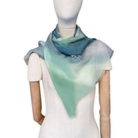Mannequin wearing a hand-painted Otta pure silk scarf on a white background in 
green and blue tones. soft, fresh and lightweight, and elegant accessory measuring 65 x 175 cm. Unique artisan scarf with hand-fringed edges, perfect for women and men.
