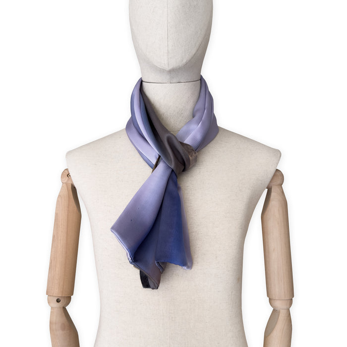 Mannequin wearing a colorful and lightweight silk scarf on a white background. Silk scarf is in shades of blue, lilac and brown colors. It is hand-painted scarf in measures 171 x 24 cm by otta-italy scarves brand.
