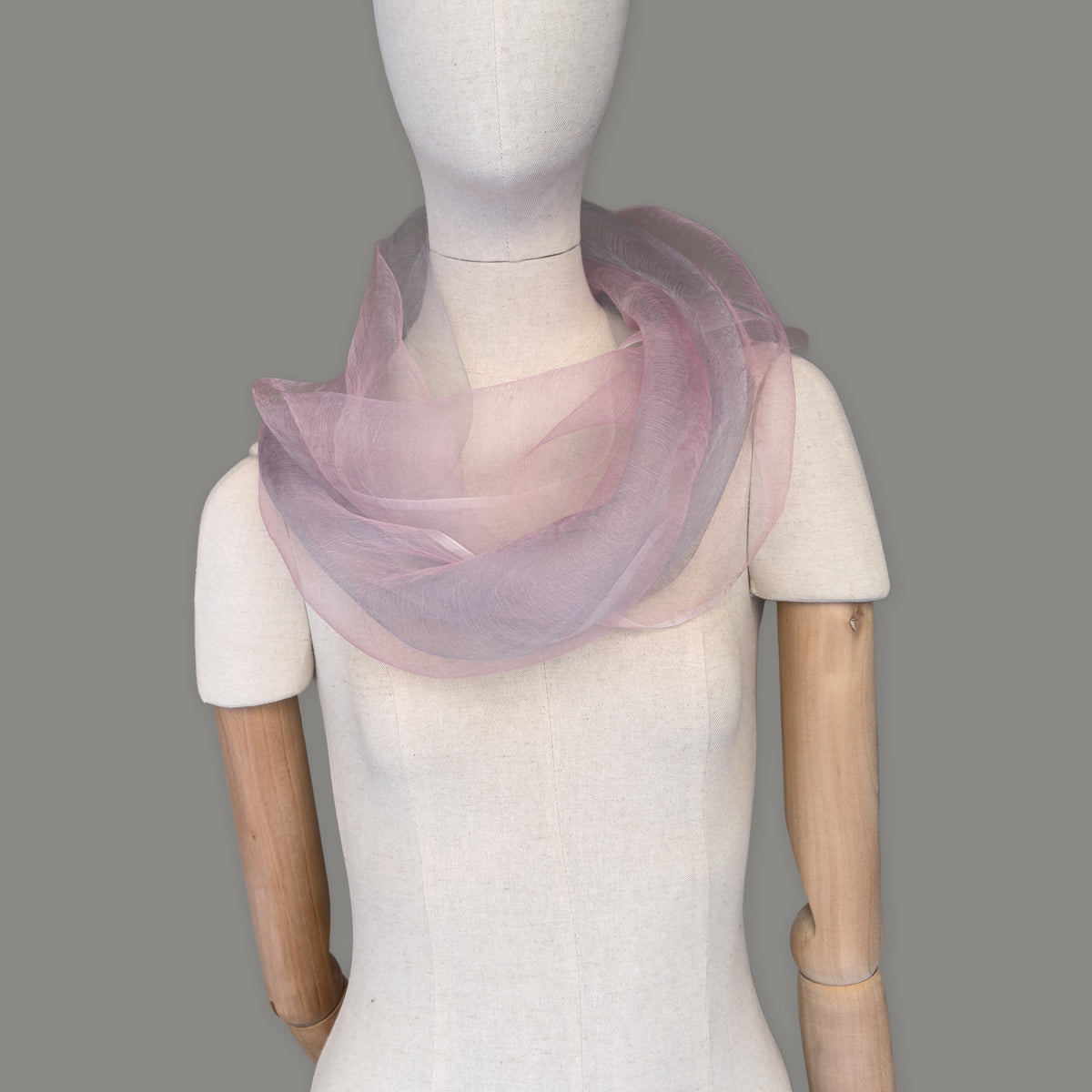 Mannequin wearing a hand-painted Otta silk scarf on a gray background in 
pink and light gray tones. Lightweight, soft, and elegant accessory measuring 50 x 200 cm. Unique artisan scarf with hand-fringed edges, perfect for women and men.
