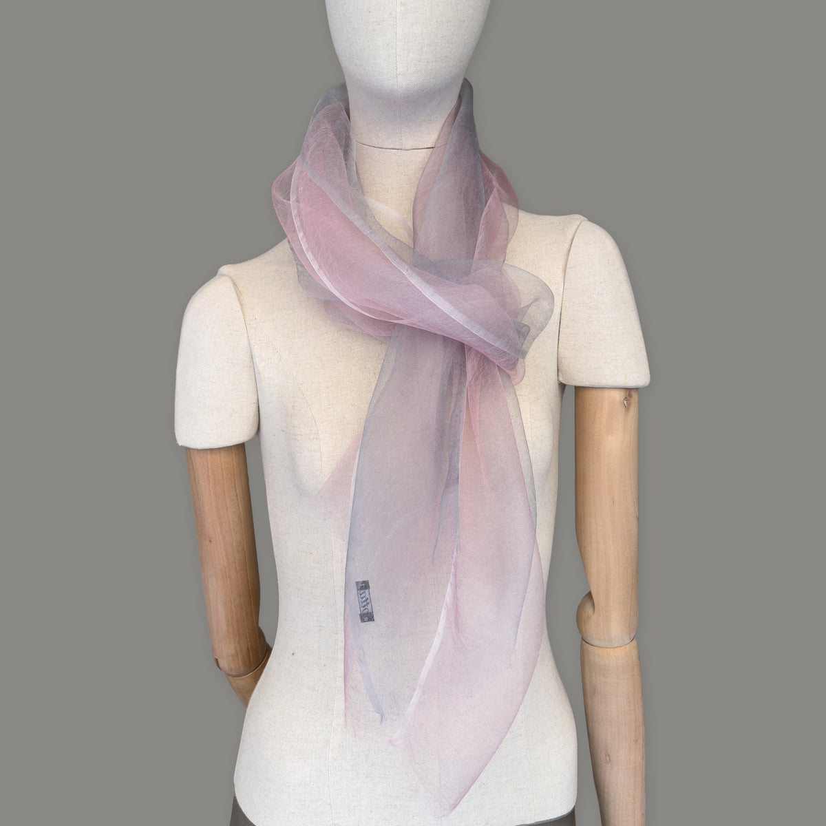 Mannequin wearing a hand-painted Otta silk scarf on a gray background in 
pink and light gray tones. Lightweight, soft, and elegant accessory measuring 50 x 200 cm. Unique artisan scarf with hand-fringed edges, perfect for women and men.
