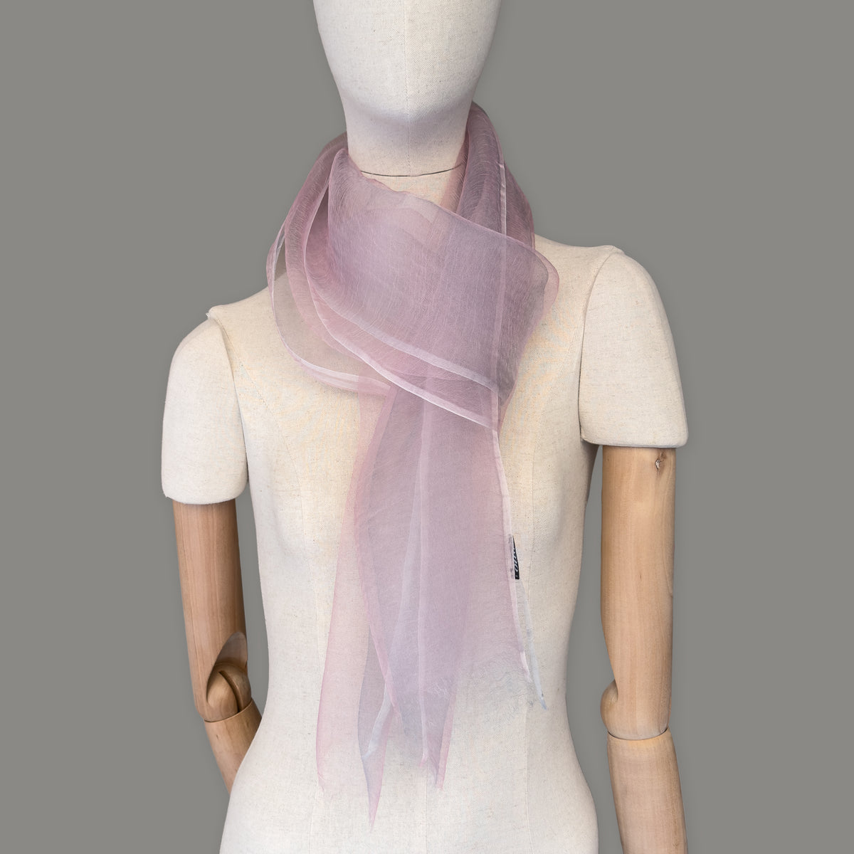 Mannequin wearing a hand-painted Otta silk scarf on a gray background in 
pink and light gray tones. Lightweight, soft, and elegant accessory measuring 50 x 200 cm. Unique artisan scarf with hand-fringed edges, perfect for women and men.
