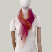 Mannequin wearing an elegant hand-painted Otta pure silk organza scarf on a gray background. Orange, red and pink tones.
