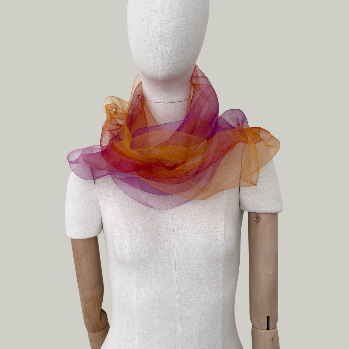 Mannequin wearing an elegant hand-painted Otta pure silk organza scarf on a gray background. Orange, red and pink tones.
