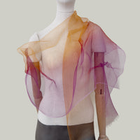 Mannequin wearing an elegant hand-painted Otta pure silk organza scarf on a gray background. Orange, red and pink tones.
