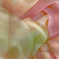 Close-up of a hand-painted Otta pure silk scarf draped softly, showcasing gentle shades of 
coral, peach, yellow and light green. Lightweight artisan accessory with delicate hand-fringed edges, measuring 65 x 175 cm.
