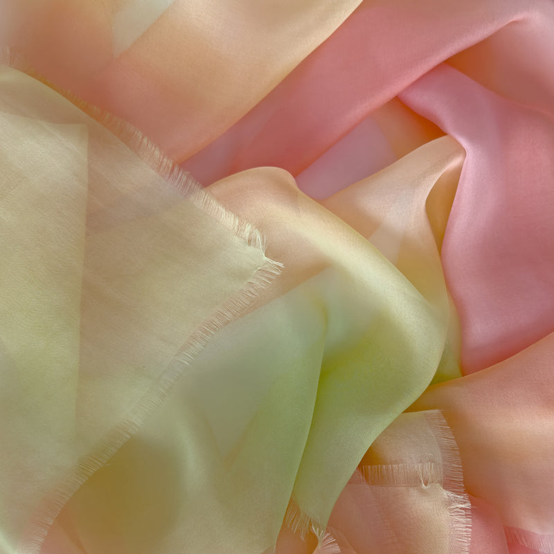Close-up of a hand-painted Otta pure silk scarf draped softly, showcasing gentle shades of 
coral, peach, yellow and light green. Lightweight artisan accessory with delicate hand-fringed edges, measuring 65 x 175 cm.
