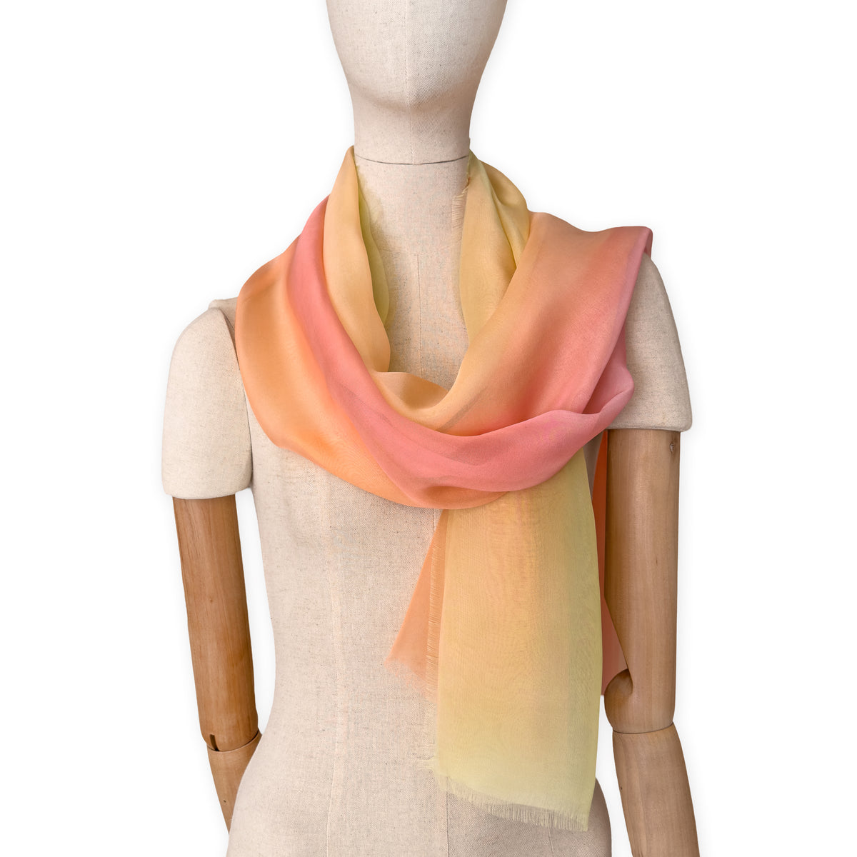 Mannequin wearing a hand-painted Otta pure silk scarf on a white background in 
coral, peach, yellow and light green tones. soft, fresh and lightweight, and elegant accessory measuring 65 x 175 cm. Unique artisan scarf with hand-fringed edges, perfect for women and men.
