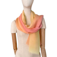 Mannequin wearing a hand-painted Otta pure silk scarf on a white background in 
coral, peach, yellow and light green tones. soft, fresh and lightweight, and elegant accessory measuring 65 x 175 cm. Unique artisan scarf with hand-fringed edges, perfect for women and men.
