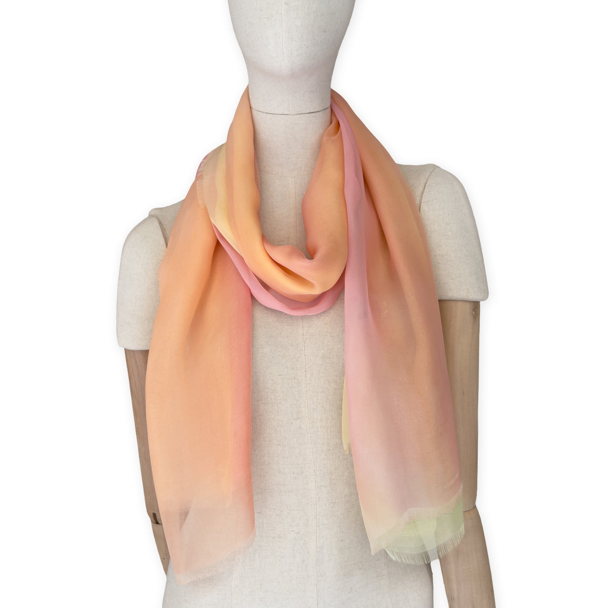 Mannequin wearing a hand-painted Otta pure silk scarf on a white background in 
coral, peach, yellow and light green tones. soft, fresh and lightweight, and elegant accessory measuring 65 x 175 cm. Unique artisan scarf with hand-fringed edges, perfect for women and men.
