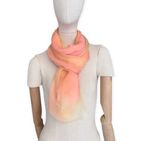Mannequin wearing a hand-painted Otta pure silk scarf on a white background in 
coral, peach, yellow and light green tones. soft, fresh and lightweight, and elegant accessory measuring 65 x 175 cm. Unique artisan scarf with hand-fringed edges, perfect for women and men.

