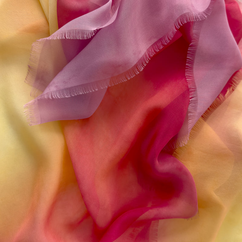 Close-up of a hand-painted Otta pure silk scarf draped softly, showcasing gentle shades of 
red, orange, fuchsia and yellow. Lightweight artisan accessory with delicate hand-fringed edges, measuring 65 x 175 cm.
