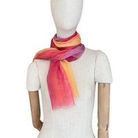 Mannequin wearing a hand-painted Otta pure silk scarf on a white background in 
red, orange, fuchsia and yellow tones. soft, fresh and lightweight, and elegant accessory measuring 65 x 175 cm. Unique artisan scarf with hand-fringed edges, perfect for women and men.
