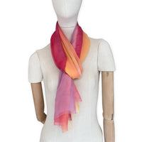 Mannequin wearing a hand-painted Otta pure silk scarf on a white background in 
red, orange, fuchsia and yellow tones. soft, fresh and lightweight, and elegant accessory measuring 65 x 175 cm. Unique artisan scarf with hand-fringed edges, perfect for women and men.
