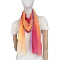 Mannequin wearing a hand-painted Otta pure silk scarf on a white background in 
red, orange, fuchsia and yellow tones. soft, fresh and lightweight, and elegant accessory measuring 65 x 175 cm. Unique artisan scarf with hand-fringed edges, perfect for women and men.
