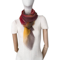 Mannequin wearing a colorful and lightweight silk scarf on a white background. Silk scarf is in shades of red, orange, yellow and brown colors. It is hand-painted scarf in measures 176 x 64 cm by otta-italy scarves brand.

