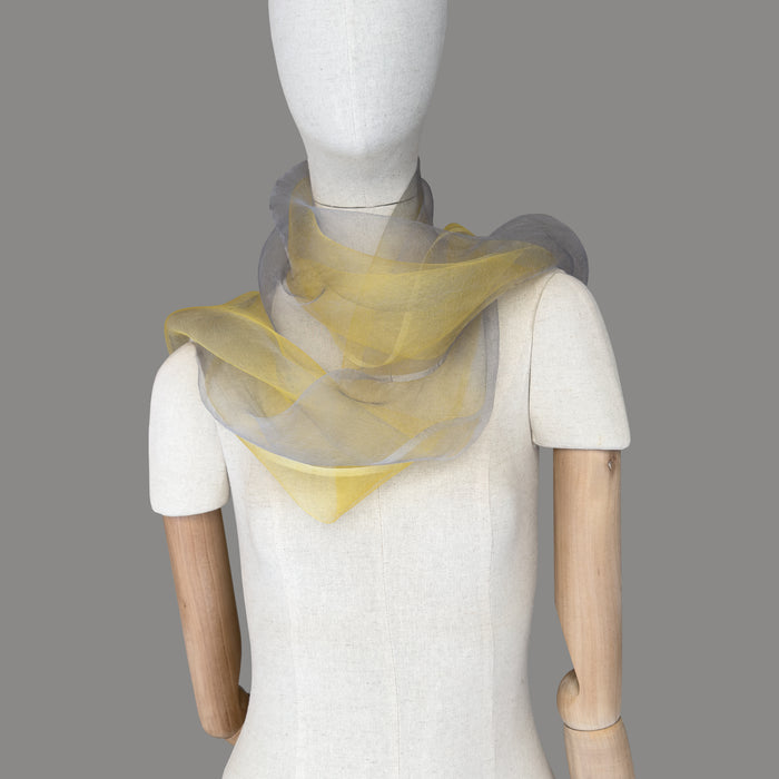 Mannequin wearing a colorful and lightweight silk scarf on a graybackground. Silk scarf is in shades of yellow and gray colors. It is hand-painted scarf in measures 50 x 194 cm by otta-italy scarves brand.
