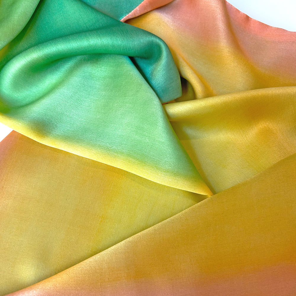 Close-up of a hand-painted Otta silk scarf draped softly, showcasing gentle shades of 
yellow, orange and green. Lightweight artisan accessory with delicate hand-hem, measuring 22 x 170 cm.

