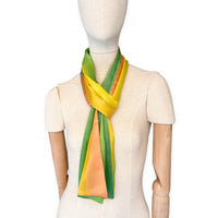 Mannequin wearing a hand-painted Otta silk scarf on a gray background in 
yellow, orange and green tones. Lightweight, soft, and elegant accessory measuring 50 x 200 cm. Unique artisan scarf with hand-hem edges, perfect for women and men.
