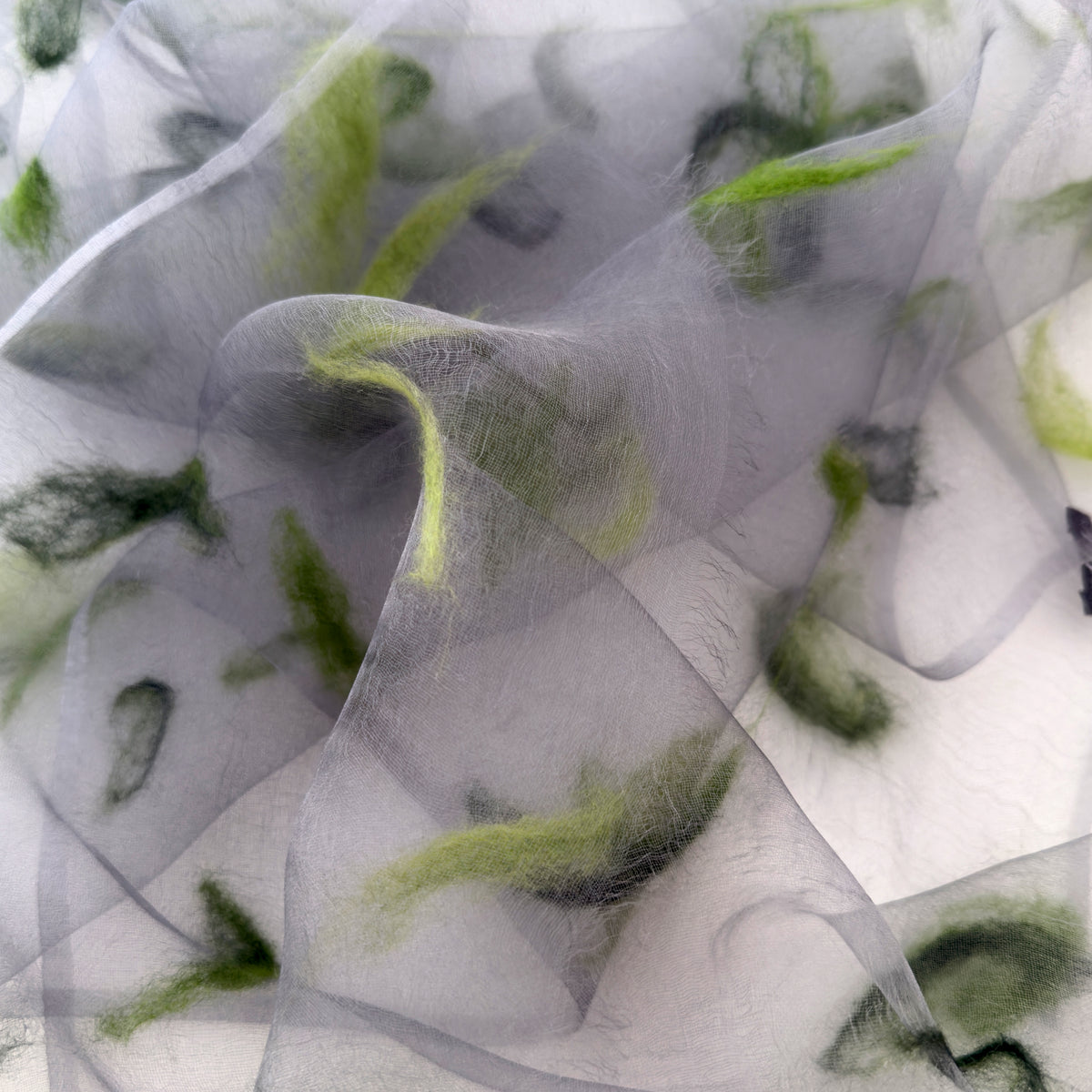 Close-up of a hand-painted Otta-Italy silk organza scarf showcasing rich shades of green hand-needle wool felting over a sheer gray background.