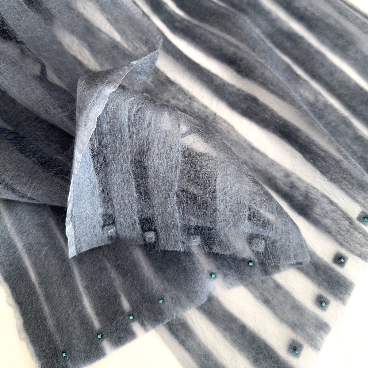 Close-up of a hand-painted Otta pure silk scarf draped softly with hand-needle felted wool showcasing gentle shades of 
gray. Lightweight artisan accessory with delicate hand-fringed edges, measuring 40 x 1