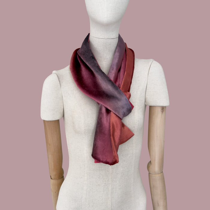 Mannequin wearing a colorful velvet scarf on a pink background. Velvet scarf is in shades of red and purple colors. It is hand-painted scarf in measures 184 x 17 cm by otta-italy scarves brand.
