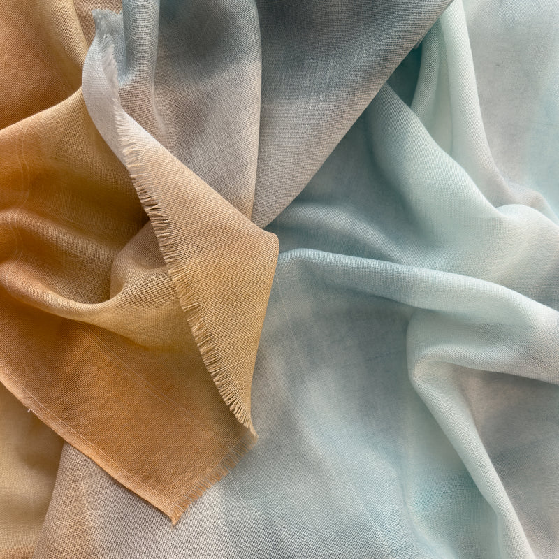 Close-up of a hand-painted Otta wool scarf draped softly, showcasing gentle shades of 
blue, gray and ocher. Lightweight artisan accessory with delicate hand-fringed edges, measuring 61 x 197 cm.