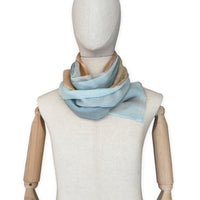 Mannequin wearing a hand-painted Otta wool scarf on a white background in 
blue, gray and ocher tones. Lightweight, warm, and elegant accessory measuring 61 x 197 cm. Unique artisan scarf with hand-fringed edges, perfect for women and men.
