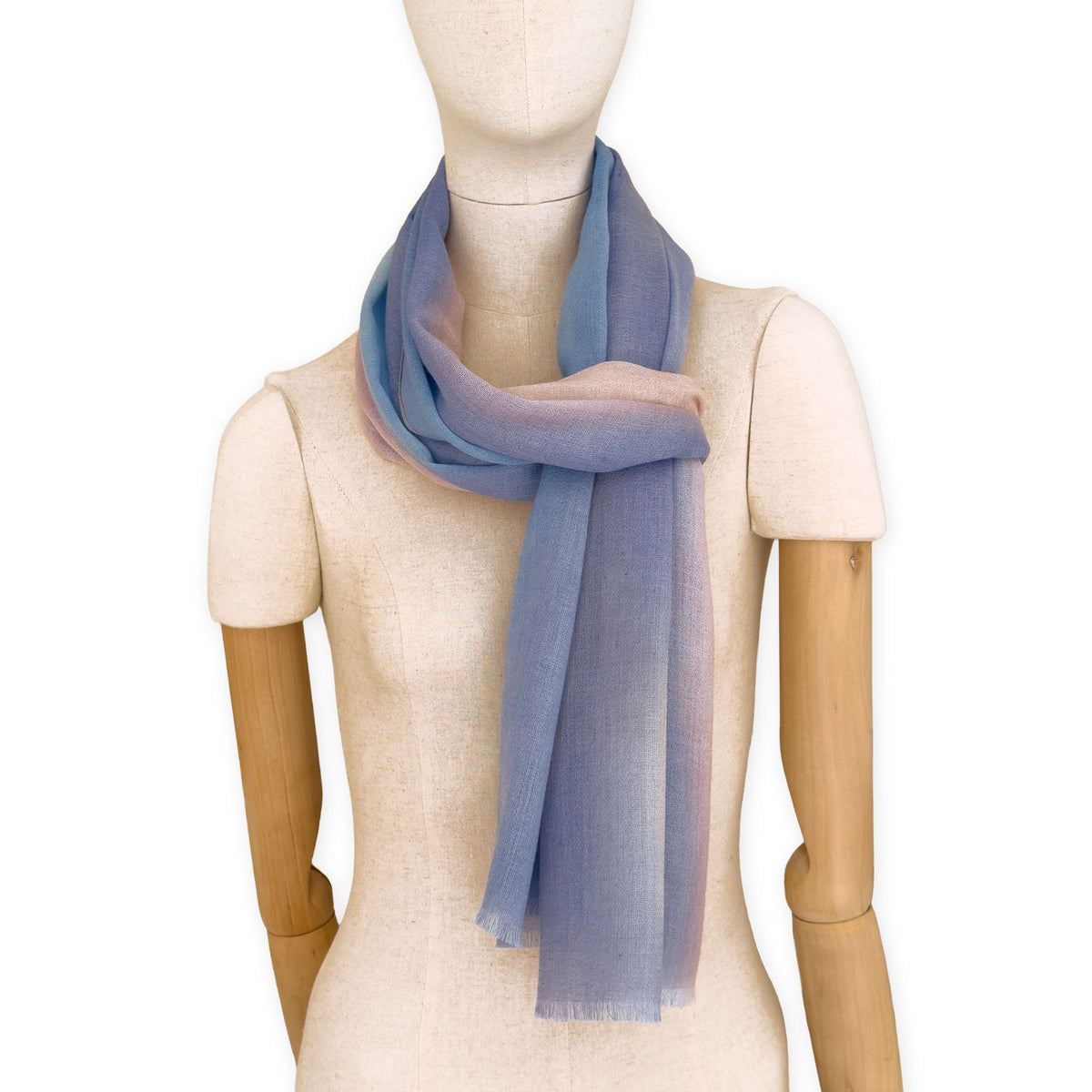 Mannequin wearing an elegant hand-painted Otta pure wool scarf on a white background. Warm blue, pink, beige and gray tones.
