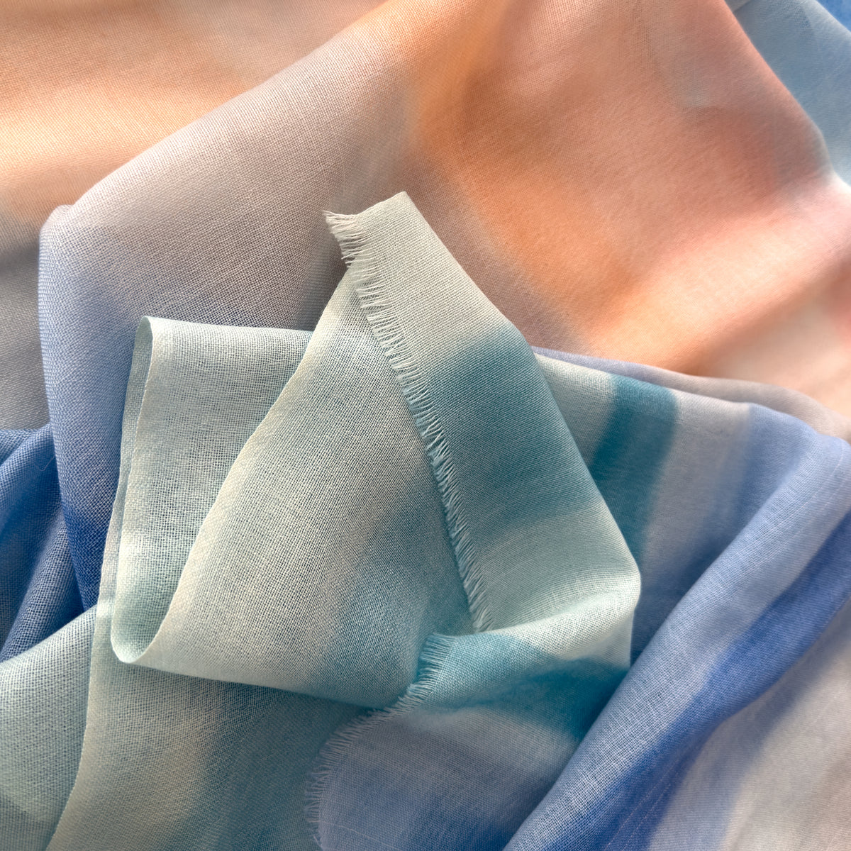 Close-up of a hand-painted Otta wool scarf draped softly, showcasing gentle shades of 
pastel blue and pink. Lightweight artisan accessory with delicate hand-fringed edges, measuring 61 x 197 cm.
