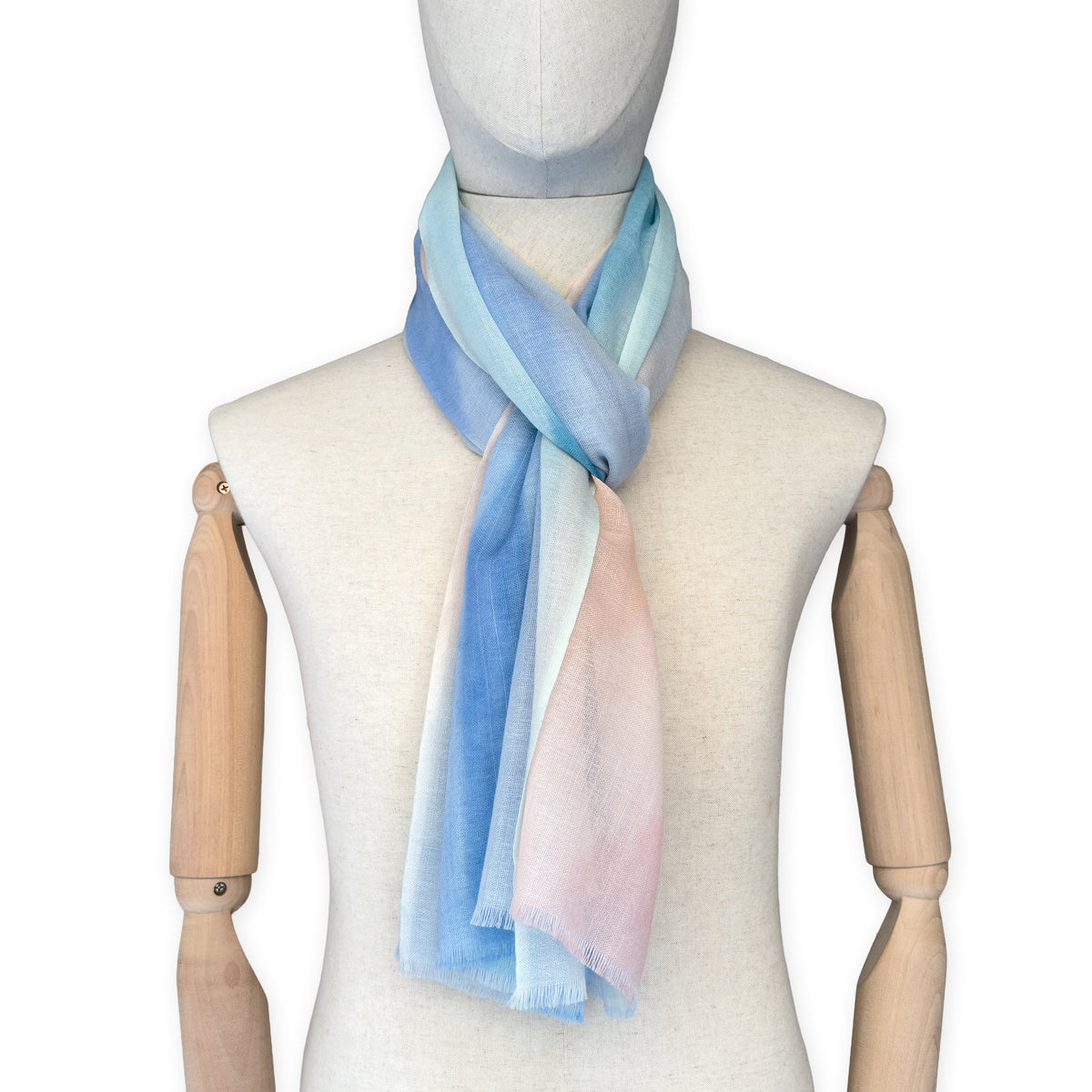 Mannequin wearing a hand-painted Otta wool scarf on a white background in 
pastel blue and pink tones. Lightweight, warm, and elegant accessory measuring 61 x 197 cm. Unique artisan scarf with hand-fringed edges, perfect for women and men.
