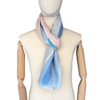 Mannequin wearing a hand-painted Otta wool scarf on a white background in 
pastel blue and pink tones. Lightweight, warm, and elegant accessory measuring 61 x 197 cm. Unique artisan scarf with hand-fringed edges, perfect for women and men.
