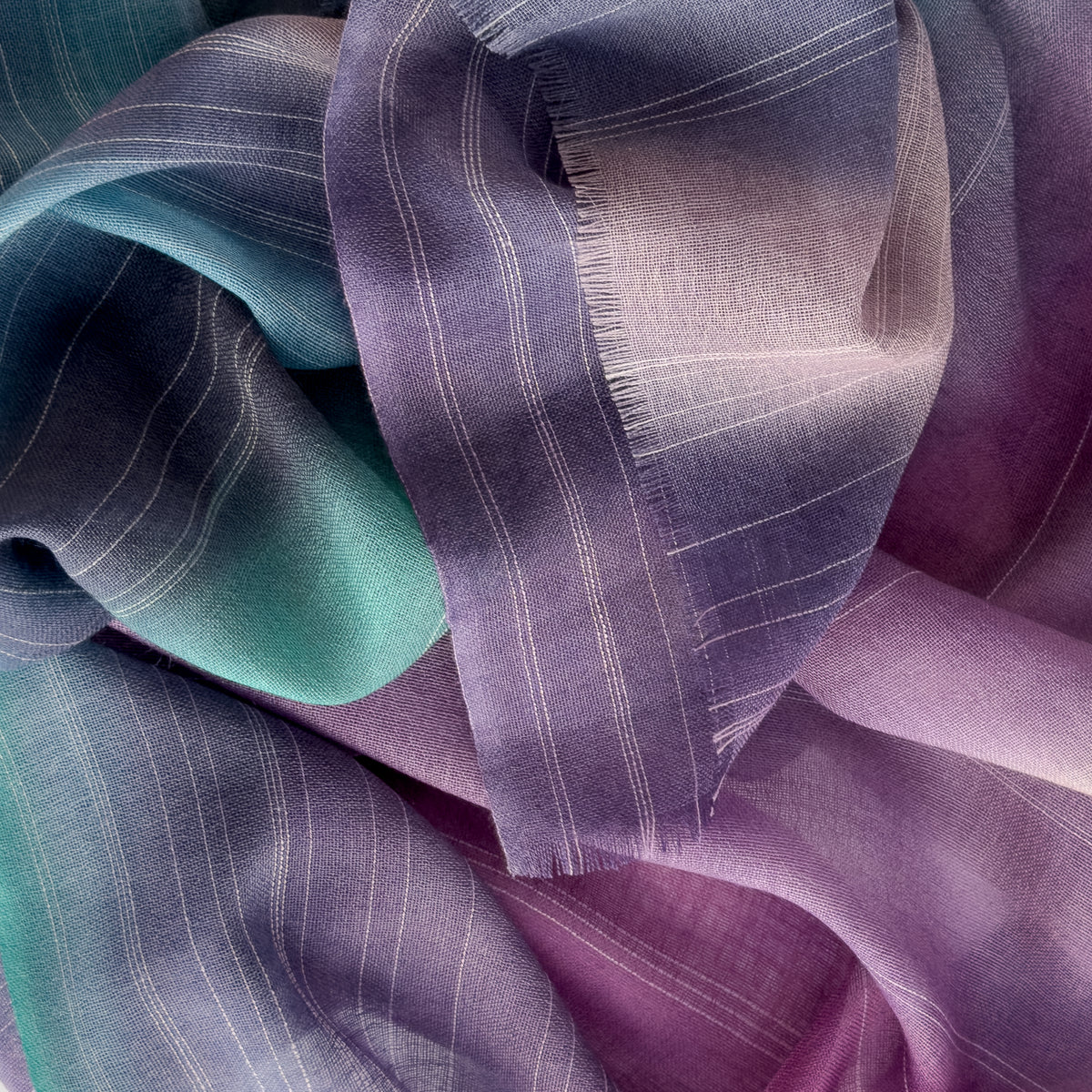 Close-up of a hand-painted Otta wool scarf draped softly, showcasing gentle shades of 
purple, emerald green and light blue. Lightweight artisan accessory with delicate hand-fringed edges, measuring 61 x 197 cm.
