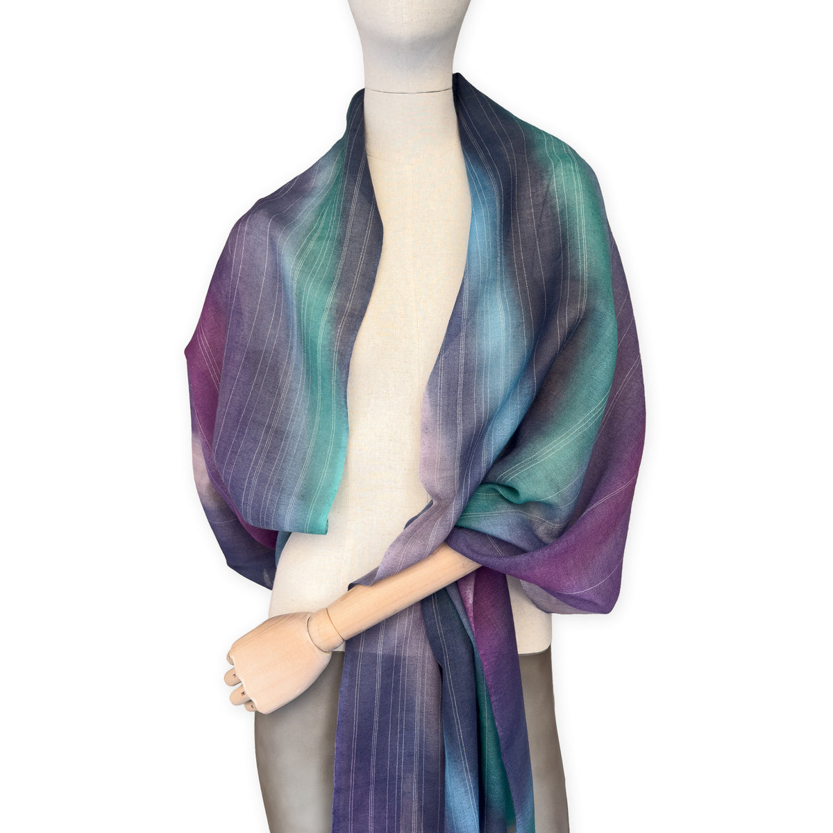 Mannequin wearing a hand-painted Otta wool scarf on a white background in 
purple, emerald green and light blue tones. Lightweight, warm, and elegant accessory measuring 61 x 197 cm. Unique artisan scarf with hand-fringed edges, perfect for women and men.
