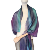 Mannequin wearing a hand-painted Otta wool scarf on a white background in 
purple, emerald green and light blue tones. Lightweight, warm, and elegant accessory measuring 61 x 197 cm. Unique artisan scarf with hand-fringed edges, perfect for women and men.
