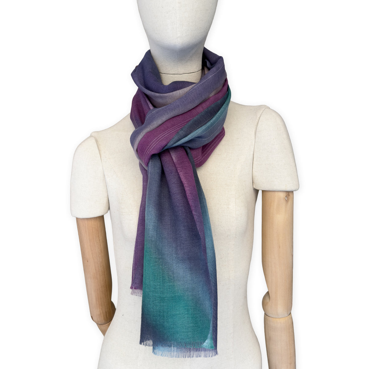 Mannequin wearing a hand-painted Otta wool scarf on a white background in 
purple, emerald green and light blue tones. Lightweight, warm, and elegant accessory measuring 61 x 197 cm. Unique artisan scarf with hand-fringed edges, perfect for women and men.
