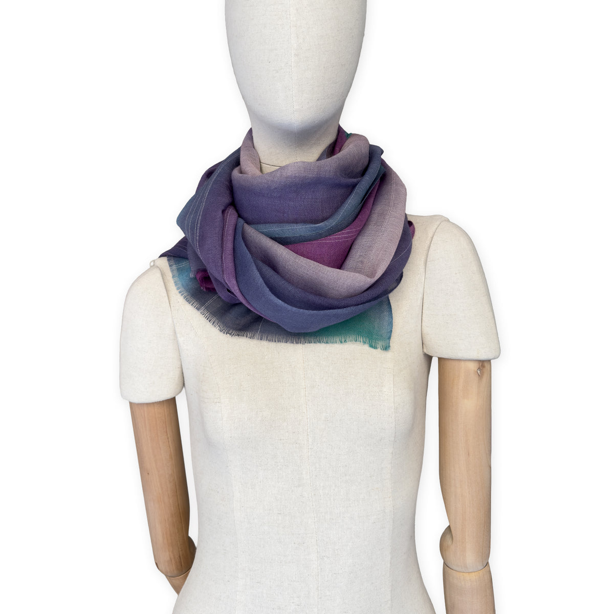 Mannequin wearing a hand-painted Otta wool scarf on a white background in 
purple, emerald green and light blue tones. Lightweight, warm, and elegant accessory measuring 61 x 197 cm. Unique artisan scarf with hand-fringed edges, perfect for women and men.
