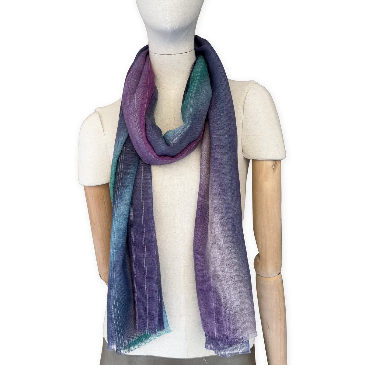 Mannequin wearing a hand-painted Otta wool scarf on a white background in 
purple, emerald green and light blue tones. Lightweight, warm, and elegant accessory measuring 61 x 197 cm. Unique artisan scarf with hand-fringed edges, perfect for women and men.
