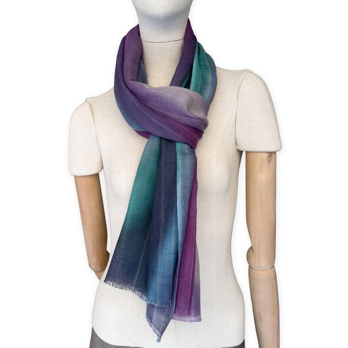 Mannequin wearing a hand-painted Otta wool scarf on a white background in 
purple, emerald green and light blue tones. Lightweight, warm, and elegant accessory measuring 61 x 197 cm. Unique artisan scarf with hand-fringed edges, perfect for women and men.
