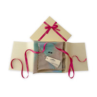 Luxury gift box with a pink ribbon containing a hand-painted Otta wool silk blend scarf on a white background.
