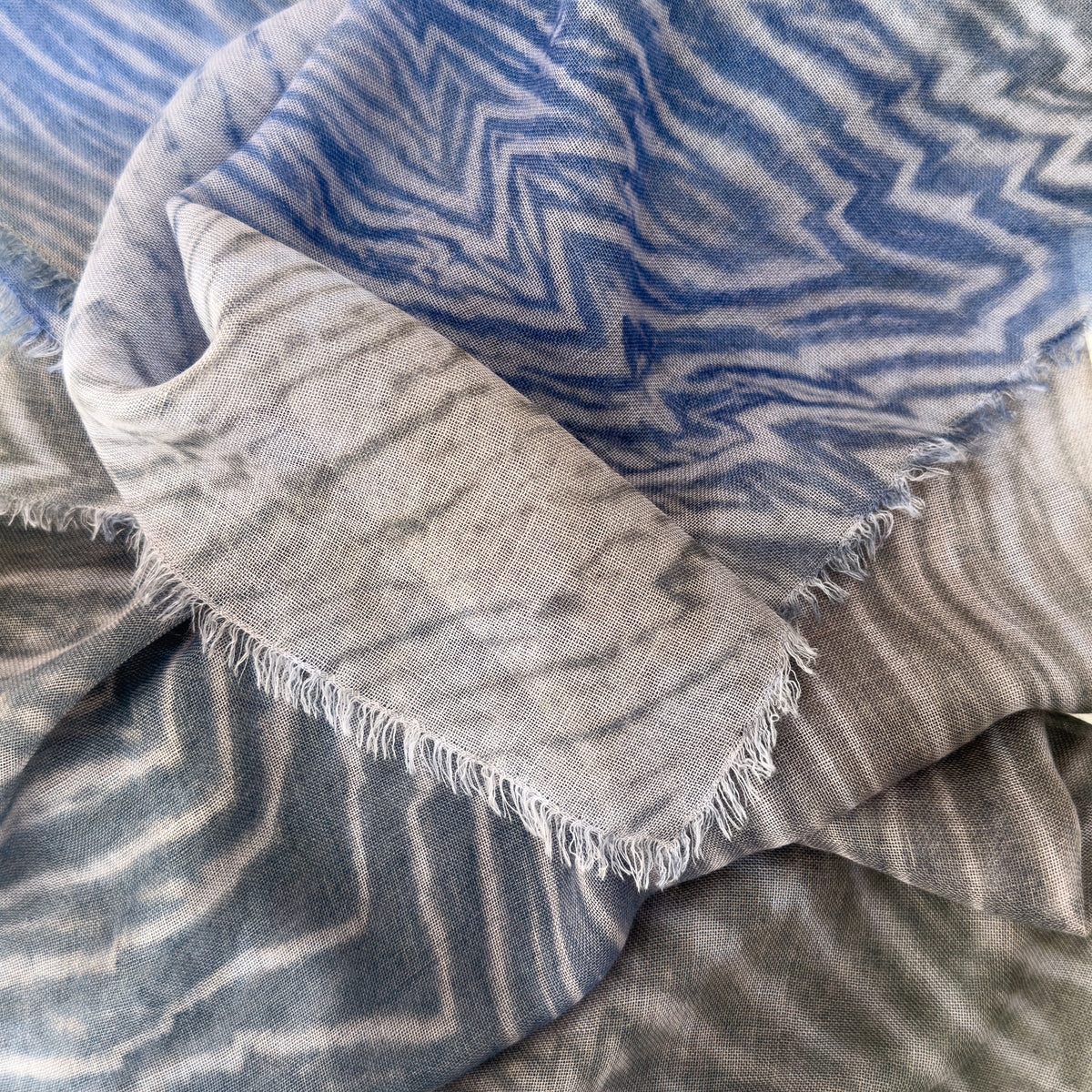 Close-up of a hand-painted Otta wool silk blend scarf showcasing soft shades of blue, gray and delicate hand-fringed edges.
