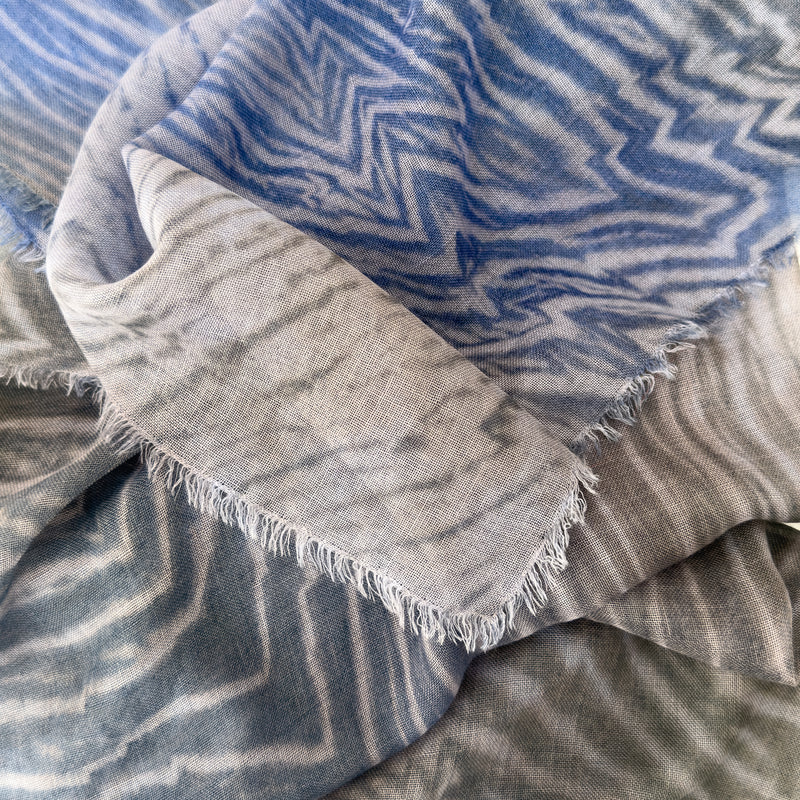 Close-up of a hand-painted Otta wool silk blend scarf showcasing soft shades of blue, gray and delicate hand-fringed edges.
