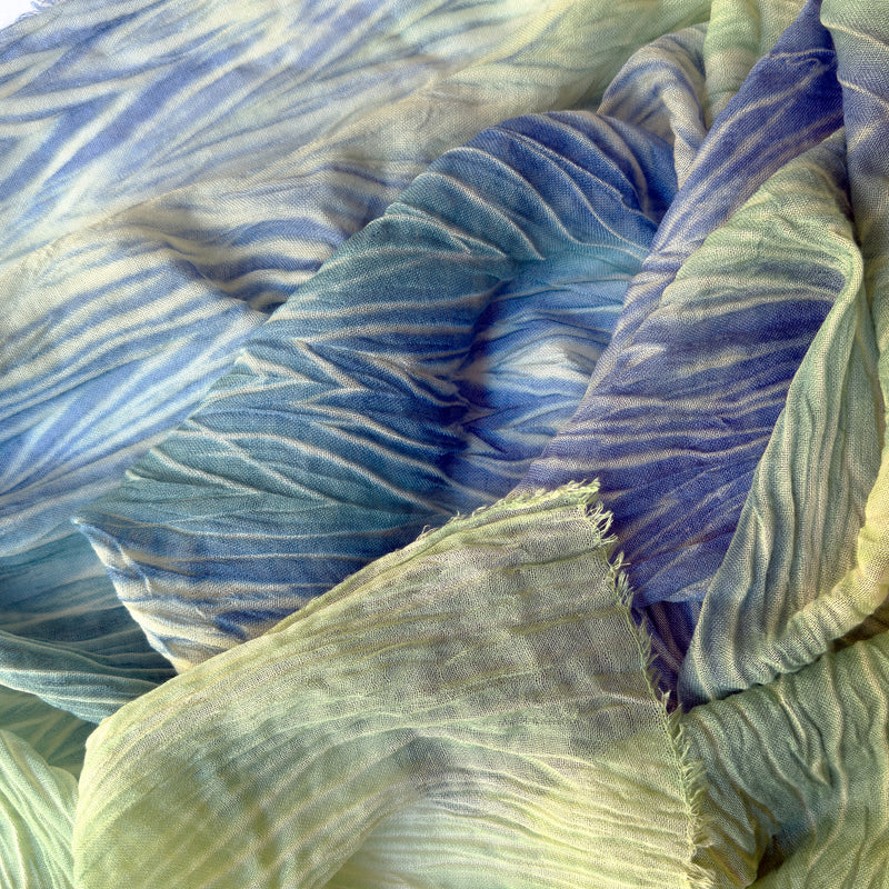 Close-up of a hand-painted Otta wool & silk scarf draped softly, showcasing gentle shades of 
blue, light green and yellow. Lightweight artisan accessory with delicate hand-fringed edges, measuring 54 x 193 cm.
