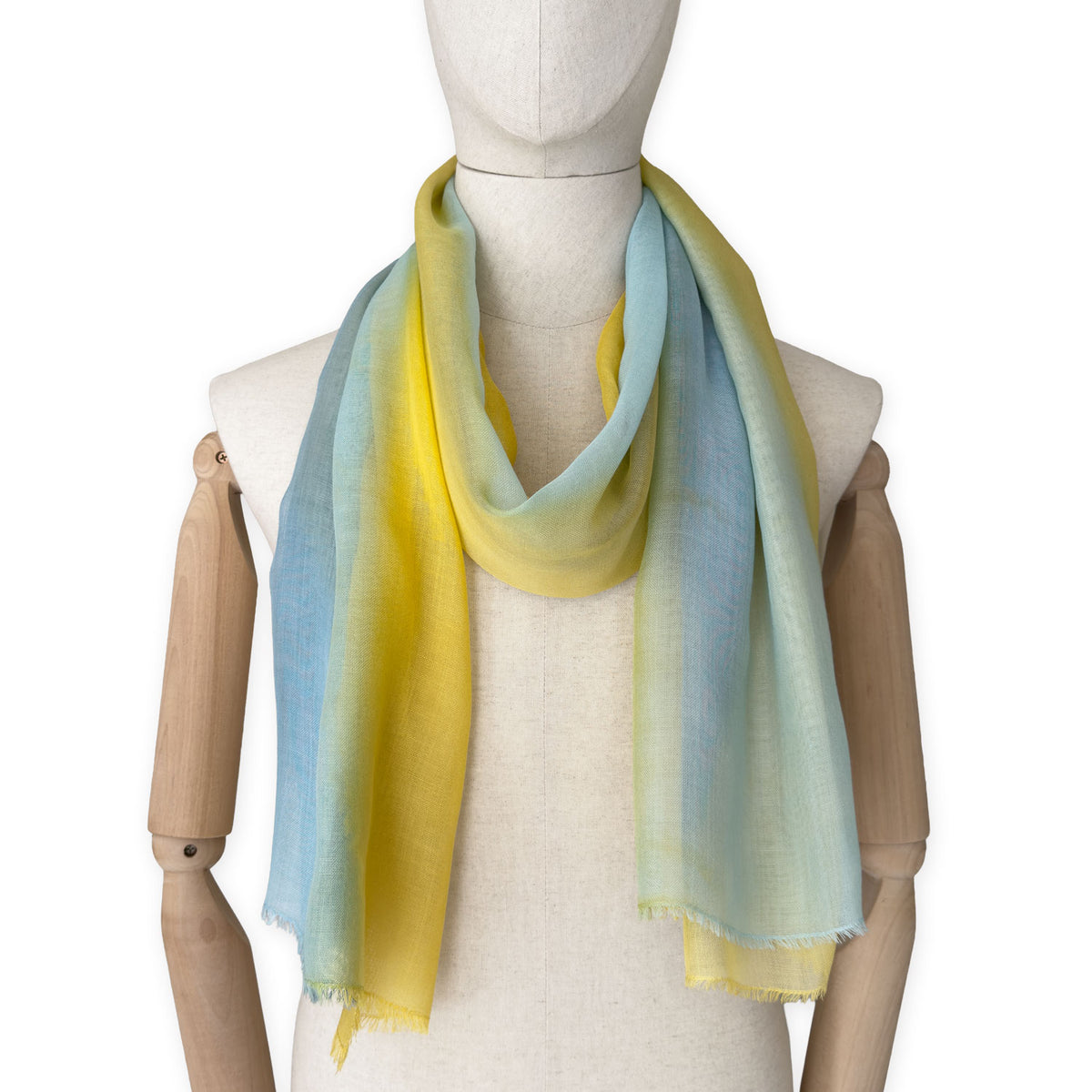 Mannequin wearing an elegant hand-painted Otta wool silk blend scarf on a white background. Warm blue and yellow tones.
