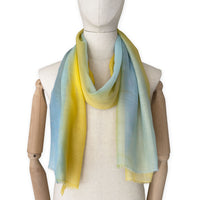 Mannequin wearing an elegant hand-painted Otta wool silk blend scarf on a white background. Warm blue and yellow tones.
