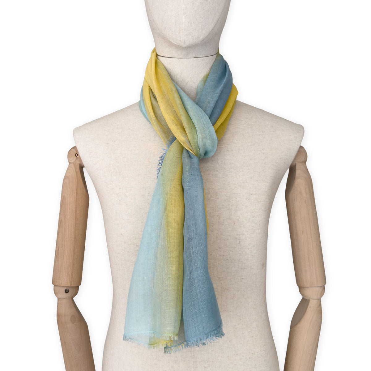 Mannequin wearing an elegant hand-painted Otta wool silk blend scarf on a white background. Warm blue and yellow tones.
