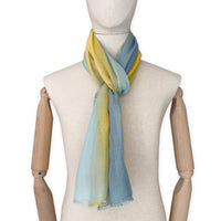 Mannequin wearing an elegant hand-painted Otta wool silk blend scarf on a white background. Warm blue and yellow tones.
