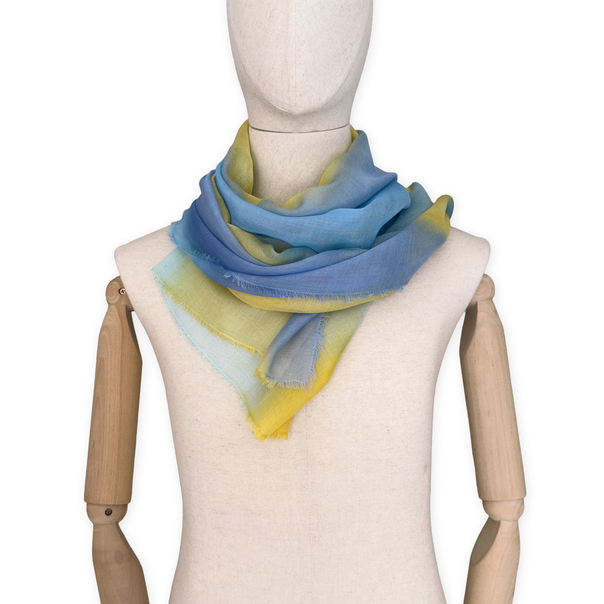 Mannequin wearing an elegant hand-painted Otta wool silk blend scarf on a white background. Warm blue and yellow tones.
