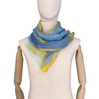 Mannequin wearing an elegant hand-painted Otta wool silk blend scarf on a white background. Warm blue and yellow tones.
