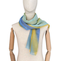 Mannequin wearing an elegant hand-painted Otta wool silk blend scarf on a white background. Warm blue and yellow tones.
