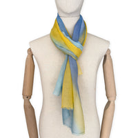 Mannequin wearing an elegant hand-painted Otta wool silk blend scarf on a white background. Warm blue and yellow tones.
